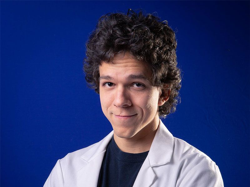 A man wearing a blue t-shirt, a white shirt over it, with a blue background.